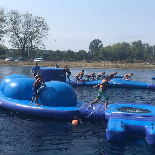 Children play on inflatable water obstacles in a lake, climbing and jumping with a sunny sky in the background.