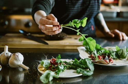 A hand reaches for fresh herbs while preparing a meal, with garlic and plates of vegetables on a kitchen counter.