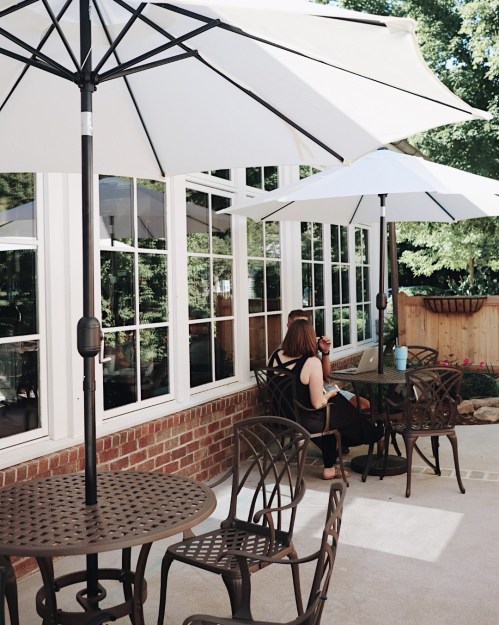 A woman sits at a table under a large umbrella, working on a laptop in a sunny outdoor patio setting.