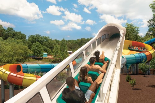 A group of people enjoying a water slide at a theme park on a sunny day, surrounded by colorful slides and greenery.