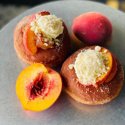 Two peach-filled donuts topped with cream and crumbled topping, alongside a whole peach and a halved peach.