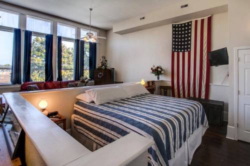 Cozy bedroom featuring a large bed, American flag, and large windows with blue curtains. Warm, inviting decor.