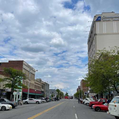 A street view featuring buildings, parked cars, and a cloudy sky, with a bank sign visible in the background.