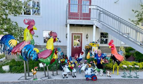 A woman stands among colorful metal rooster sculptures and various animal decorations outside a building.