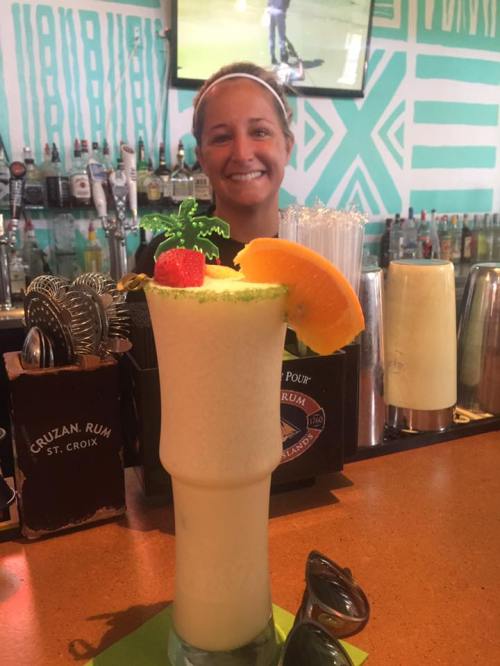 A bartender smiles behind a colorful tropical drink garnished with a strawberry and orange slice.