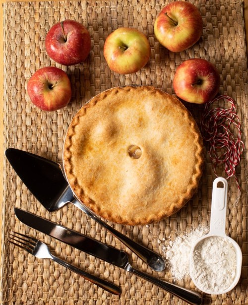 A golden apple pie surrounded by red apples, utensils, flour, and twine on a woven mat.