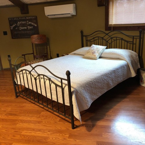 A simple metal bed frame with a white quilt in a cozy room featuring wooden floors and a chair in the background.