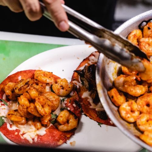 A hand using tongs to add shrimp to a plate of colorful tacos with melted cheese and garnishes.