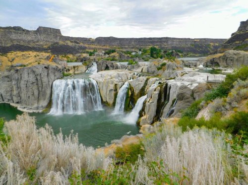 A scenic view of a waterfall cascading into a green pool, surrounded by rocky cliffs and sparse vegetation.