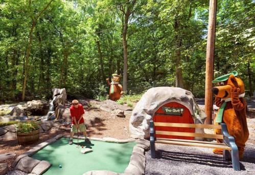 A child plays mini-golf in a wooded area, with cartoon bear characters nearby and a pro shop sign in the background.