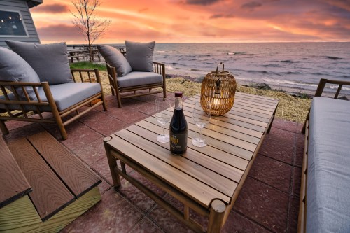 A cozy outdoor seating area by the water at sunset, featuring a table with wine and glasses, surrounded by comfortable chairs.