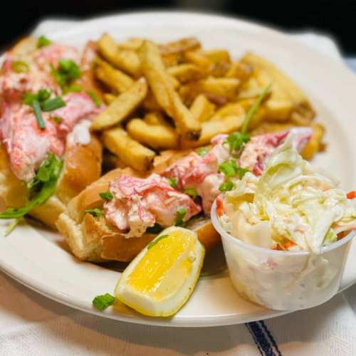 A plate featuring two lobster rolls, fries, coleslaw, and a lemon wedge.