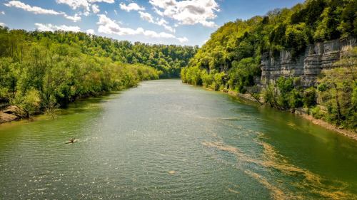 A serene river flows between lush green hills and rocky cliffs under a bright blue sky with fluffy clouds.