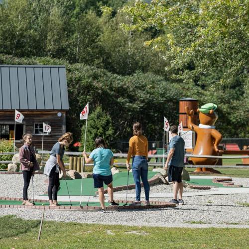 A group of five people playing mini-golf outdoors, with colorful flags and a large cartoon character in the background.