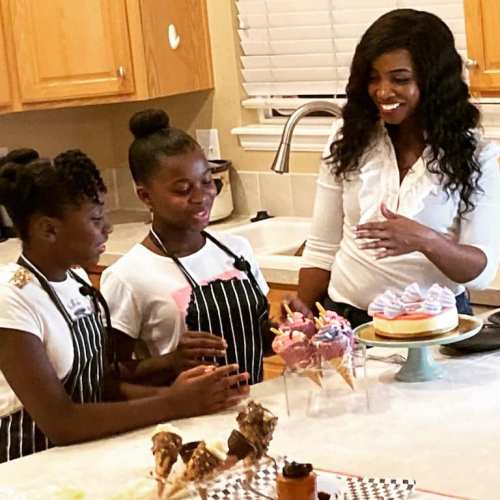 Two girls in aprons and a woman smile while decorating desserts in a kitchen. A cake and treats are on the counter.
