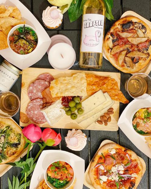 A vibrant spread of charcuterie, pizza, salads, and drinks, surrounded by flowers and candles on a wooden table.