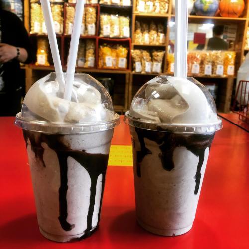 Two milkshakes with whipped cream and chocolate drizzle, served in clear cups with straws, on a red table.