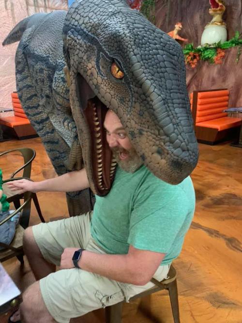 A man with a surprised expression sits at a table next to a large dinosaur sculpture with its mouth open.