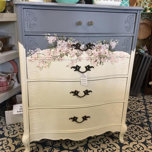 A vintage dresser with a gray and cream finish, featuring floral designs and ornate black handles.