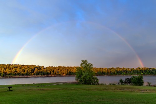 A vibrant rainbow arcs over a river, with lush green trees lining the shore under a partly cloudy sky.