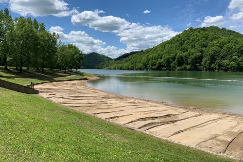 A serene lakeside view with sandy shores, green hills, and a blue sky dotted with clouds.