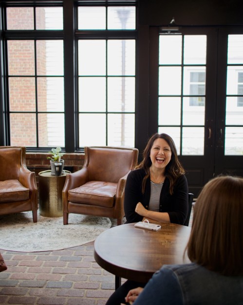 A woman laughs while sitting at a table in a cozy, well-lit room with large windows and comfortable chairs.