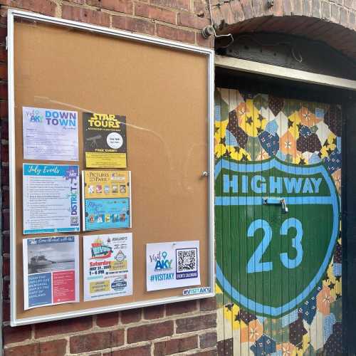 Bulletin board with event flyers and a colorful mural of "Highway 23" on a brick wall. QR code for more info.