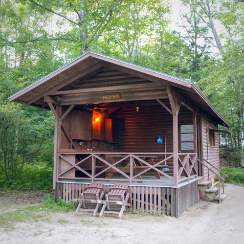 A rustic wooden cabin with a porch, surrounded by trees, featuring two chairs and a warm light glowing inside.