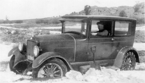A vintage car stuck in snow, with a driver inside, set against a snowy landscape.