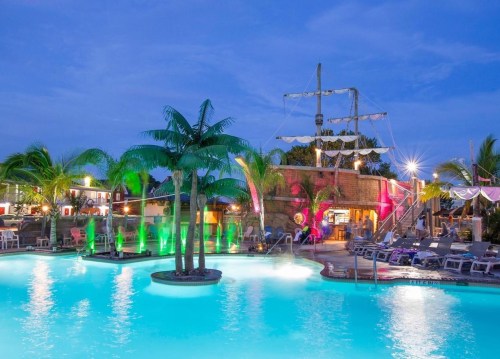 A vibrant pool area with palm trees, colorful lights, and a pirate ship structure in the background at dusk.