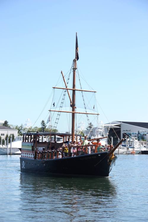 A pirate-themed ship with passengers sails in a marina on a clear day, surrounded by boats and buildings.