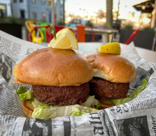 Two crispy fish sandwiches on a bed of lettuce, garnished with lemon wedges, served in a basket lined with newspaper.