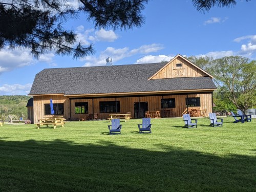 A wooden building with a sloped roof, surrounded by green grass and blue chairs, under a clear blue sky.