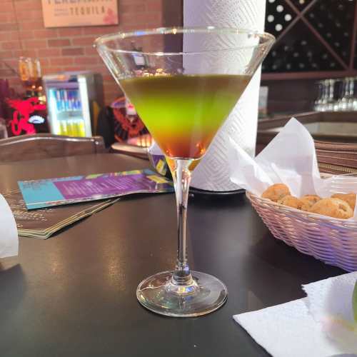 A colorful cocktail in a martini glass, featuring green and orange layers, on a bar counter with snacks nearby.