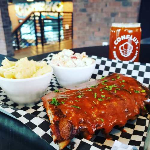 A plate of BBQ ribs with sides of macaroni and coleslaw, alongside a glass of beer on a checkered table.
