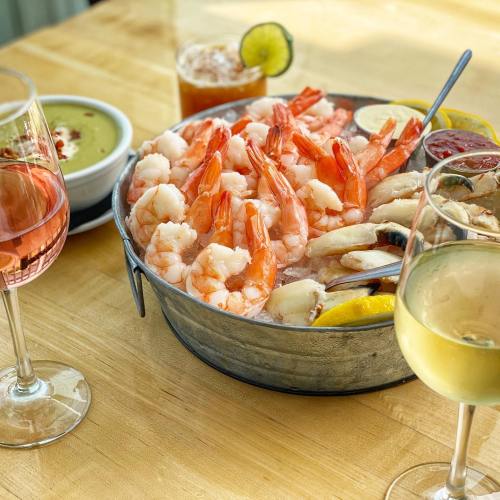 A metal bowl filled with shrimp, lemon slices, and dipping sauces, accompanied by drinks in glasses on a wooden table.