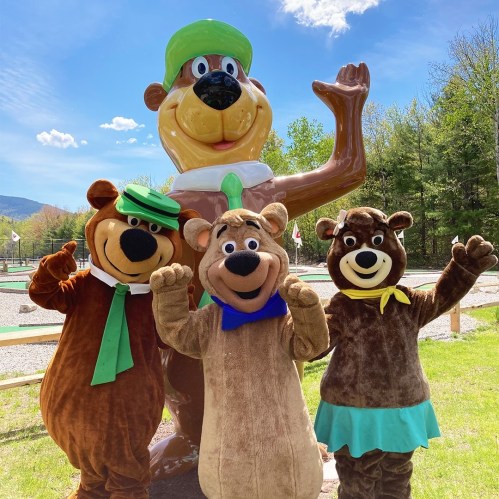 Four cartoon bear characters in costumes pose cheerfully in front of a large bear statue on a sunny day.