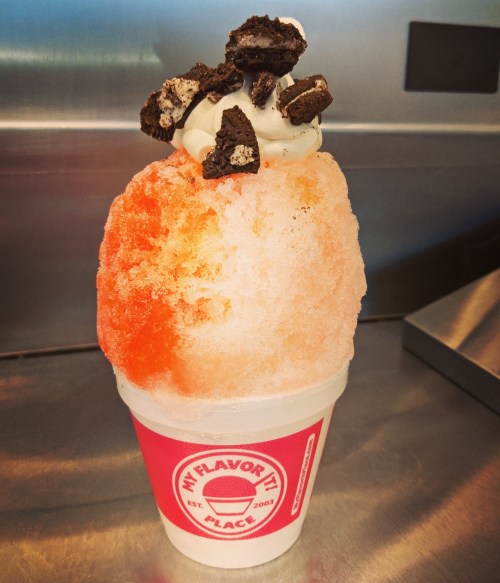 A colorful snow cone topped with whipped cream and crushed cookies, served in a branded cup.