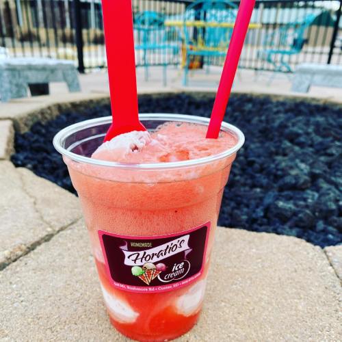 A cup of strawberry ice cream with a swirl of white, topped with two red spoons, set against a patio background.