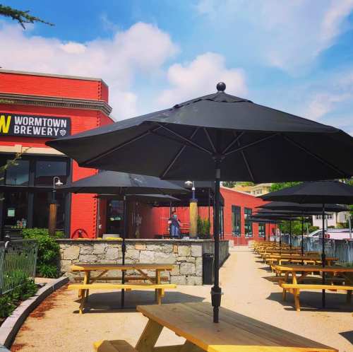 Outdoor seating area with wooden picnic tables and black umbrellas at Wormtown Brewery, under a blue sky.