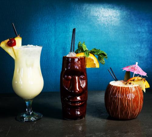 Three colorful cocktails: a creamy drink in a glass, a tiki mug with a face, and a coconut cup, all garnished with fruit.