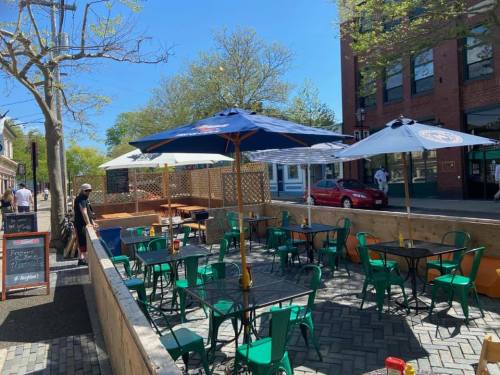 Outdoor dining area with green chairs and umbrellas, surrounded by trees and buildings on a sunny day.