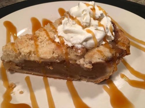 A slice of apple pie topped with whipped cream and drizzled with caramel sauce on a white plate.
