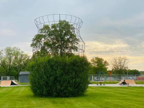 A large wire sculpture resembling a vase stands atop a bush in a park, with skate ramps and trees in the background.