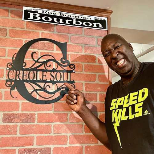 A person in a black "Speed Kills" shirt smiles and points at a decorative sign that reads "Creole Soul" on a brick wall.