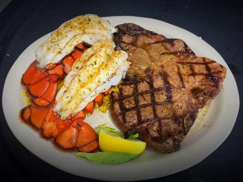 A plate featuring grilled lobster tails, a steak, and a lemon wedge, garnished with lettuce.