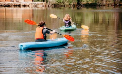 Two people kayaking on a calm lake, one in a blue kayak and the other in a green kayak, surrounded by trees.