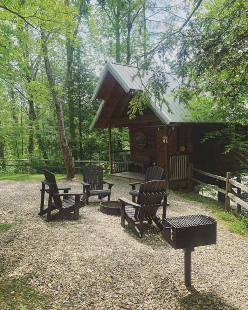 A cozy cabin surrounded by trees, featuring a gravel area with chairs and a grill.