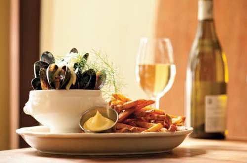 A bowl of mussels with herbs, served with sweet potato fries and a glass of white wine in the background.