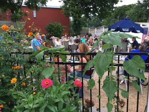 A lively outdoor gathering with people seated at tables, surrounded by flowers and greenery.